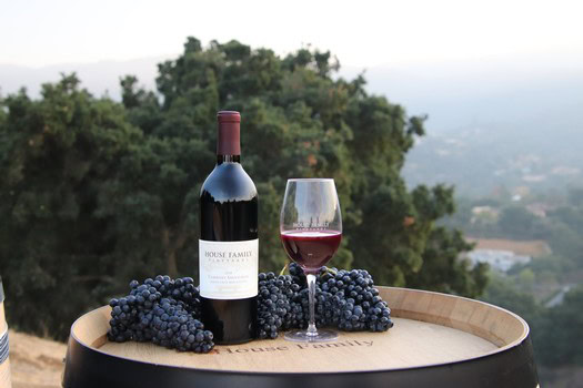 A bottle of Red Wine from House Family Vineyards, a glass, and grapes upon a wine barrel.